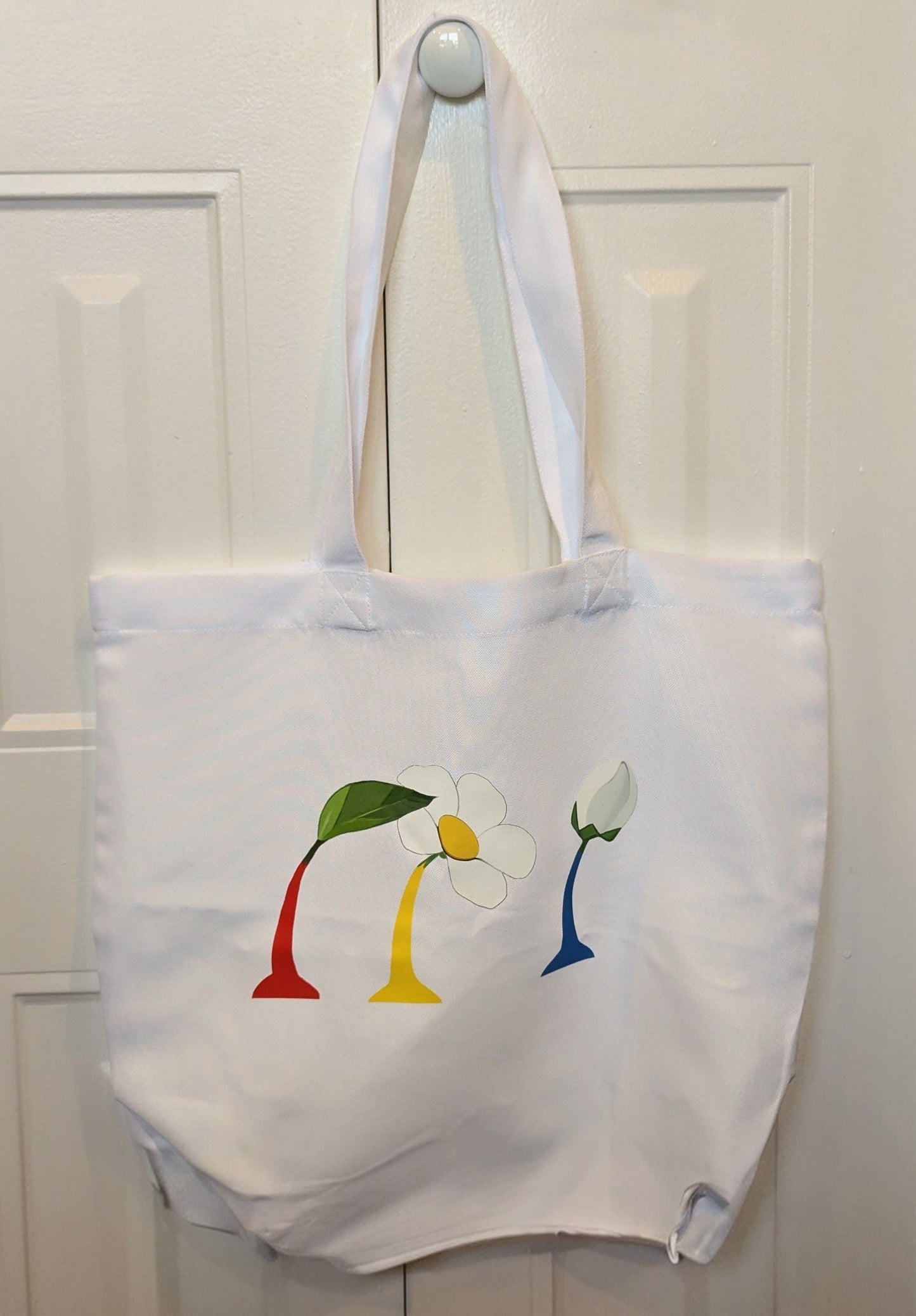 White tote bag with colorful flower design hanging on a white door.